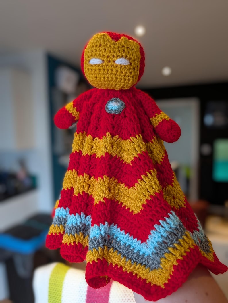 May include: A handmade crochet lovey featuring a stuffed head resembling a red and gold helmet with white eyes. The body is a soft, textured blanket with a chevron pattern in alternating stripes of red, gold, light blue, and gray yarn.