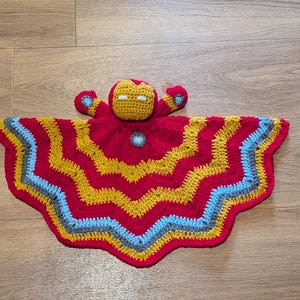 May include: A handmade crochet security blanket featuring a central character head with a red and gold mask and white eyes. The attached blanket has a wavy, chevron-style pattern in alternating bands of red, gold, light blue, and gray yarn.