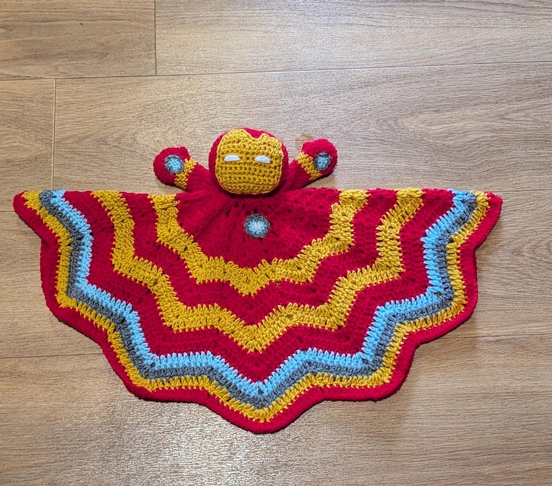 May include: A handmade crochet security blanket featuring a central character head with a red and gold mask and white eyes. The attached blanket has a wavy, chevron-style pattern in alternating bands of red, gold, light blue, and gray yarn.