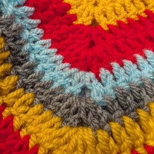 May include: A close-up of a crocheted textile featuring concentric circular rows of yarn in vibrant colors. The pattern displays alternating bands of bright red, golden yellow, light blue, and charcoal gray, showcasing the textured stitch work.