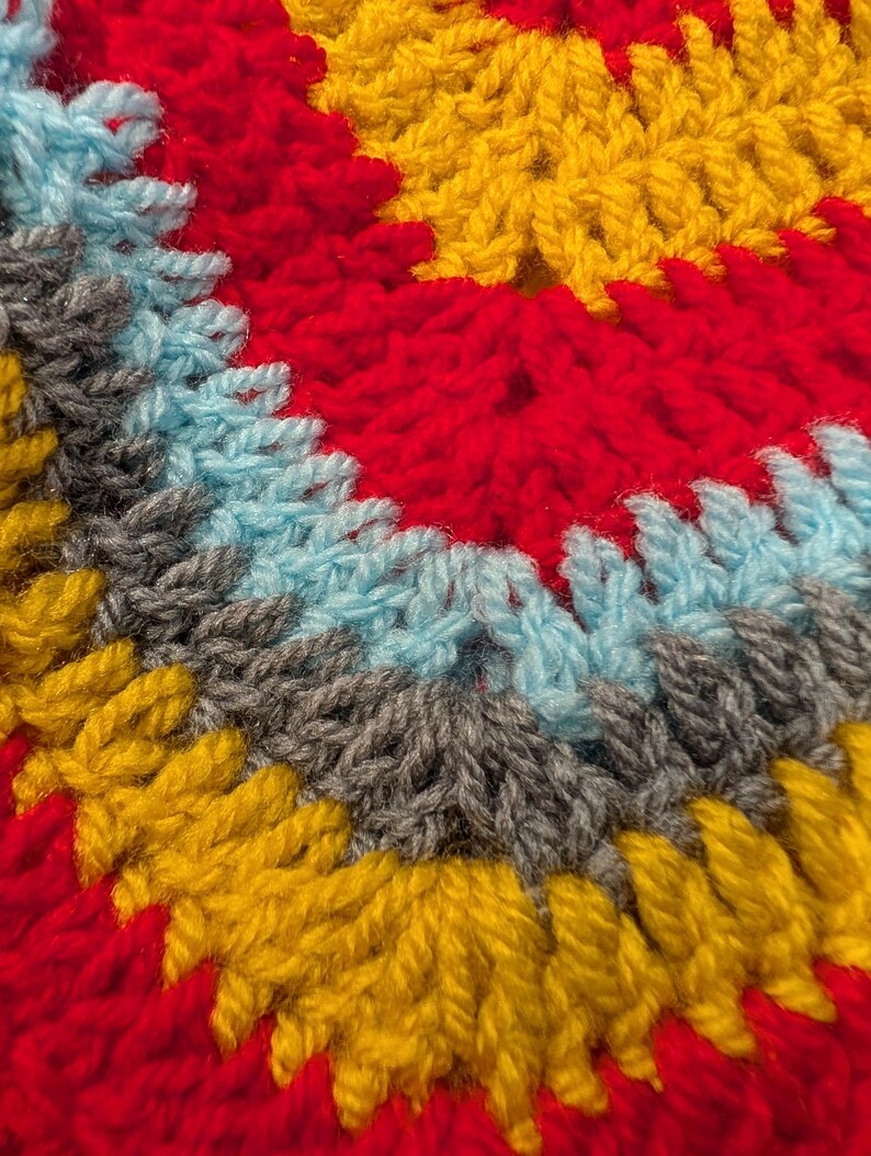 May include: A close-up of a crocheted textile featuring concentric circular rows of yarn in vibrant colors. The pattern displays alternating bands of bright red, golden yellow, light blue, and charcoal gray, showcasing the textured stitch work.
