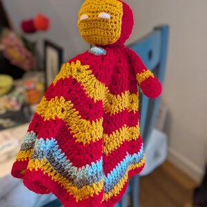 May include: A crochet security blanket featuring a stuffed head resembling a red and gold helmet with white eye slits. The attached blanket body displays a chevron pattern in alternating rows of red, gold, light blue, and grey yarn textures.