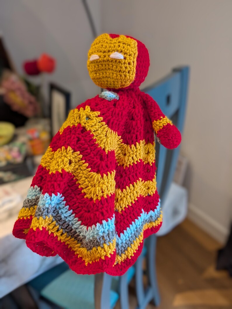 May include: A crochet security blanket featuring a stuffed head resembling a red and gold helmet with white eye slits. The attached blanket body displays a chevron pattern in alternating rows of red, gold, light blue, and grey yarn textures.