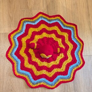 May include: A circular crochet blanket featuring a wavy, star-like pattern with concentric rings of red, mustard yellow, light blue, and gray yarn. A small, round, red crocheted plush figure with two short arms is attached to the center of the blanket.