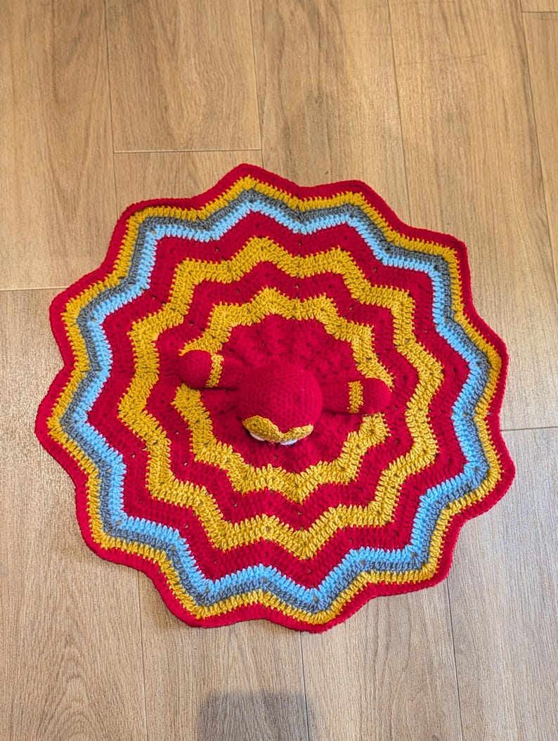 May include: A circular crochet blanket featuring a wavy, star-like pattern with concentric rings of red, mustard yellow, light blue, and gray yarn. A small, round, red crocheted plush figure with two short arms is attached to the center of the blanket.