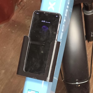 May include: A black smartphone holder mounted on the light blue frame of an electric bicycle. The phone is secured within the holder, displaying a dark screen with a purple rose graphic and the time 6:15. The bike frame features the text XP4.
