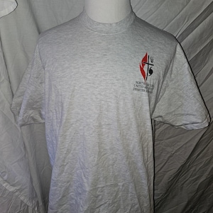 May include: A light gray heathered short-sleeve t-shirt featuring a small logo on the left chest. The logo includes a red flame, a black cross, and the text North Carolina United Methodist Disaster Response printed in black lettering below the icon.