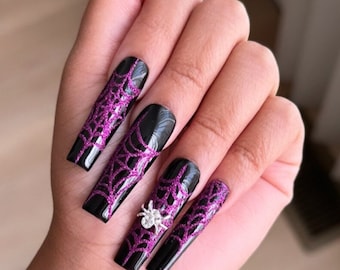 May include: Long, square-shaped artificial nails featuring a glossy black base with a vibrant purple glitter spiderweb design. One nail is accented with a small, silver-toned three-dimensional spider charm positioned over the web pattern.