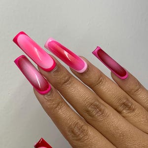May include: A hand with long, square-shaped acrylic nails. The nails feature a gradient design, transitioning from light pink to a deeper shade of pink, with a bright pink outline. The nails are a bold and stylish fashion statement.