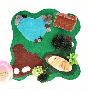Könnte beinhalten: Eine grüne Filzspielmatte mit einem Teich, einem Haus, einem Baum, einem Baumstamm und Steinen. Die Matte ist perfekt für fantasievolles Spielen.