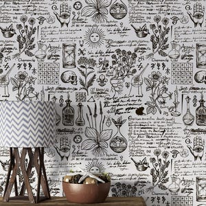May include: A wooden table with a lamp and a bowl in front of a wallpaper background. The wallpaper features a repeating pattern of black and white illustrations of plants, skulls, hourglasses, and handwritten text. The lamp has a grey and white chevron pattern.