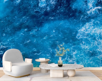 May include: A white armchair with a blue ocean wave background. The armchair is in a living room setting with a small table and a vase with green plants.