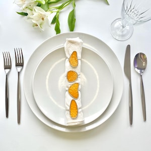 May include: A white place setting with a folded napkin adorned with four orange slice napkin rings. Silverware, including forks, a knife, and a spoon, is arranged around the plate. White flowers and a clear glass complete the elegant table setting.