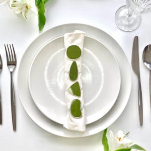 Puede incluir: Una servilleta de lino blanco doblada está adornada con tres servilleteros verde oliva en varias formas orgánicas. La servilleta descansa sobre un plato blanco, con cubiertos y un vaso visibles. El conjunto está sobre una superficie blanca.
