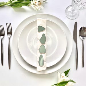 May include: A table setting with white plates, silverware, and a napkin ring. The napkin ring features four sea glass pieces in a silver setting. White flowers and a wine glass are also present, creating an elegant dining scene.