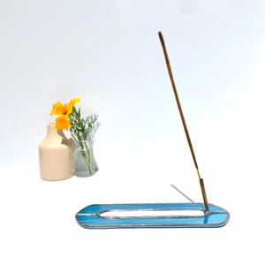Op de afbeelding: Een blauw en zilver wierookhouder met een brandende wierookstok. De houder heeft een langwerpige ovale vorm met een lichtblauw glazen ontwerp en zilveren accenten. Een kleine vaas met gele bloemen staat op de achtergrond.