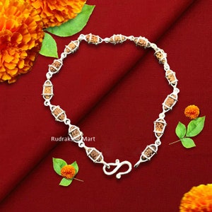May include: A silver bracelet with brown rudraksha beads. The bracelet has a hook clasp and is on a red background.