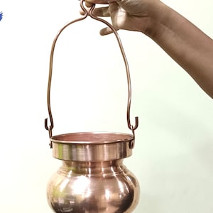 Puede incluir: Una olla de cobre colgante con fondo redondeado y borde ancho. La olla tiene un asa de cobre unida al borde.