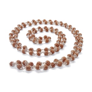 May include: A brown and silver mala necklace made of rudraksha beads and a silver chain. The necklace is laid out in a spiral shape on a white background.