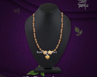May include: A brown rudraksha mala necklace with a silver chain and a heart-shaped pendant. The necklace is displayed on a black stand. The text "Rudraksha Mart" is visible below the necklace. The text "|| शिवाय गौरीवदनाब्जवृन्द सूर्याय दक्षाध्वरनाशकाय श्रीनीलकण्ठाय वृषध्वजाय तस्मै शिकाराय नमः शिवाय। ||" is visible below the stand. The text "34 शिवाय" is visible in the top right corner of the image.