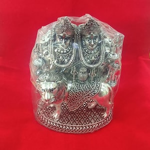 Può includere: Statua argentata di una dea indù che cavalca un leone. La dea è raffigurata con due teste ed è adornata con gioielli intricati. La statua è posta su una base circolare con un bordo decorativo.