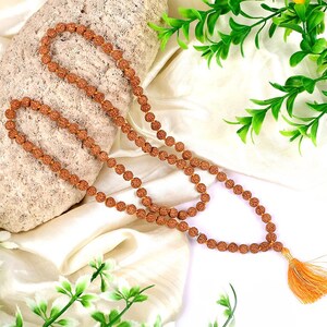 May include: A brown mala bead necklace with a tassel made of orange and gold thread. The necklace is lying on a white fabric background with green plants.