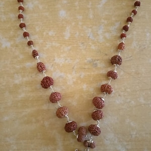 Può includere: Una collana di perline marroni con un ciondolo d'argento. Le perline sono rotonde e hanno una superficie testurizzata. Il ciondolo ha la forma di una mezzaluna con una piccola perla rotonda al centro.