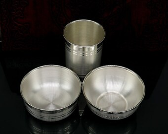 May include: Three silver drinking vessels, two bowls and one cup, with a decorative band around the top of each. The vessels are on a black surface.