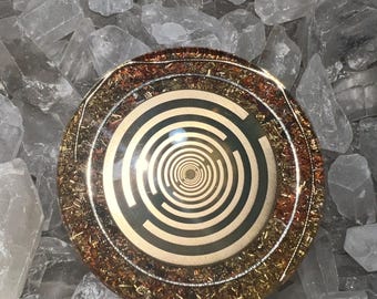 May include: A circular resin disc featuring a central gold-toned spiral maze pattern on a dark background, surrounded by a wide border of embedded copper and gold-colored metallic shavings, all encased in a smooth, glossy, transparent finish.