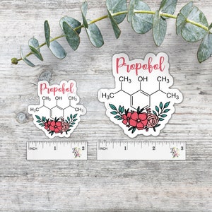 May include: Two white stickers with floral designs and the chemical formula for Propofol. The stickers are on a wooden surface.