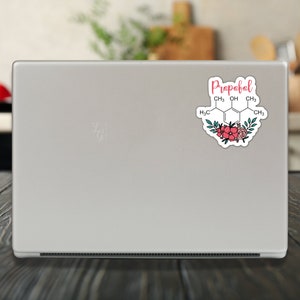 May include: A white laptop with a sticker on the back. The sticker features a chemical structure with the word "Propofol" written above it. The chemical structure is surrounded by pink and green flowers.