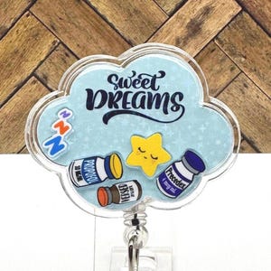 以下が含まれることがあります： 黒い文字で「Sweet Dreams」と書かれた青い雲の形をしたバッジホルダー。バッジホルダーには顔のある星と、「Propolol」、「Versed」、「Precedex」と書かれたラベルの付いた小さなボトルが3つあります。