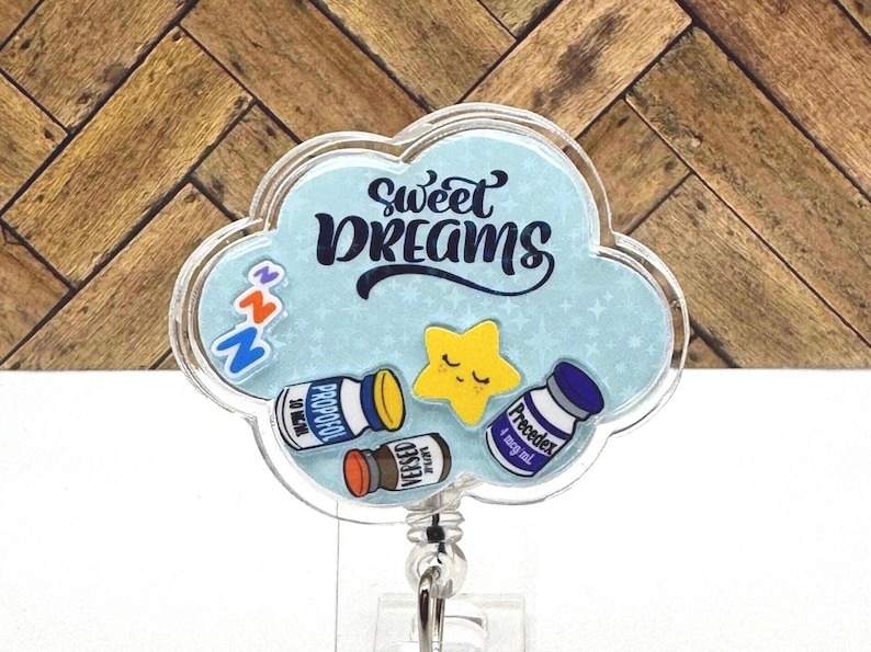 以下が含まれることがあります： 黒い文字で「Sweet Dreams」と書かれた青い雲の形をしたバッジホルダー。バッジホルダーには顔のある星と、「Propolol」、「Versed」、「Precedex」と書かれたラベルの付いた小さなボトルが3つあります。