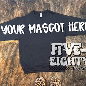 Può includere: Una felpa blu navy a maniche lunghe con la scritta "YOUR MASCOT HERE" in bianco sul petto e sulle maniche. Il design include "FIVE-EIGHTY designs" in un font retrò. Lo sfondo è una superficie a motivi marrone e nero.