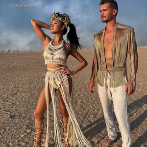 May include: A couple wearing white and gold fringe clothing poses in a desert setting. The woman wears a headpiece with gold accents and a long white fringe skirt. The man wears a white shirt and a gold fringe vest. The background is a sandy desert with a blue sky.