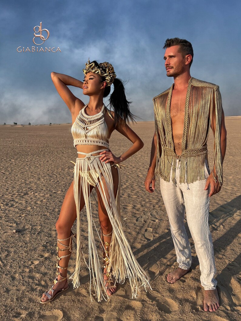 May include: A couple wearing white and gold fringe clothing poses in a desert setting. The woman wears a headpiece with gold accents and a long white fringe skirt. The man wears a white shirt and a gold fringe vest. The background is a sandy desert with a blue sky.
