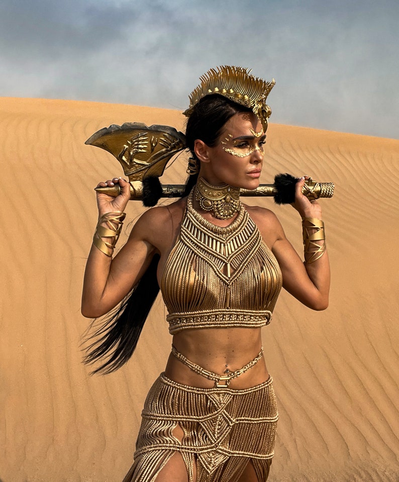 May include: A woman wearing a gold-colored outfit with a matching headpiece and holding a gold-colored axe. She is standing in a desert setting.