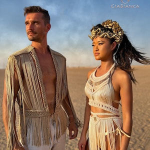 May include: Two people wearing gold and cream crocheted clothing stand in a desert setting. The woman is wearing a headpiece with gold accents. The text "Giabianca" is visible in the upper right corner of the image.