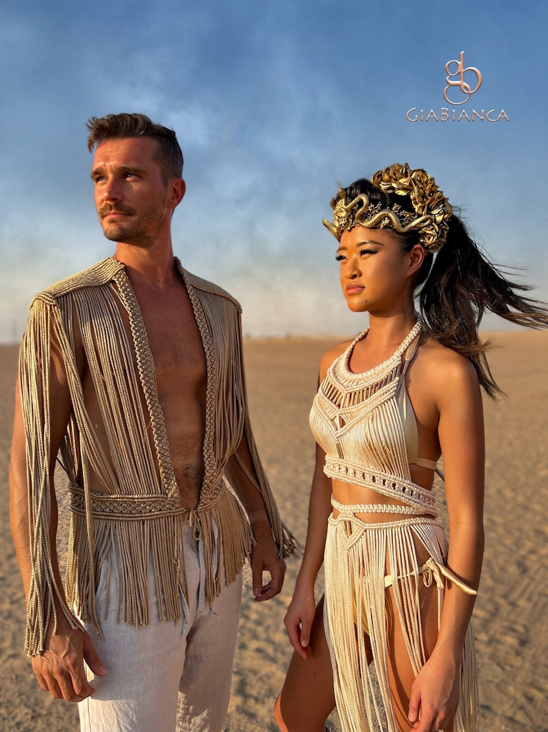 May include: Two people wearing gold and cream crocheted clothing stand in a desert setting. The woman is wearing a headpiece with gold accents. The text "Giabianca" is visible in the upper right corner of the image.