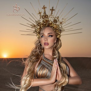 May include: A woman wearing a gold crown with a floral design and a gold top with a criss-cross pattern. The crown has a halo of spikes and crosses. The woman is standing in a desert setting with a sunset in the background.