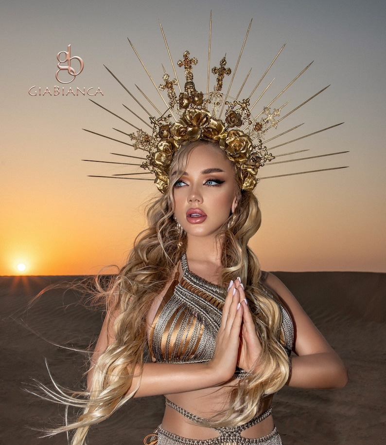 May include: A woman wearing a gold crown with a floral design and a gold top with a criss-cross pattern. The crown has a halo of spikes and crosses. The woman is standing in a desert setting with a sunset in the background.