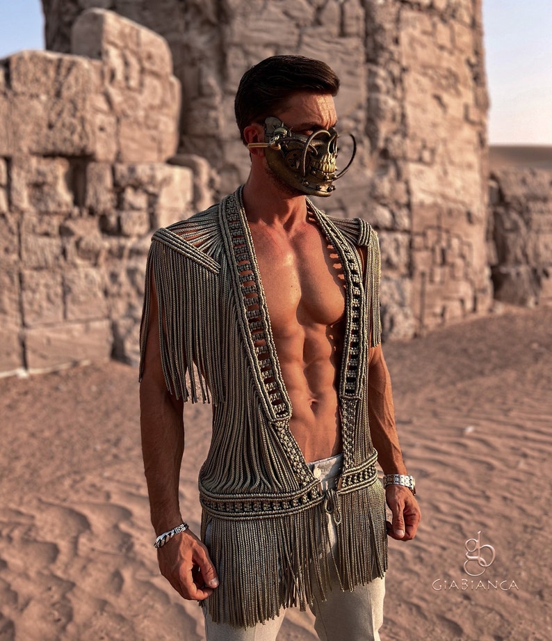 May include: A person wearing a  fringed,  beige  vest with a  skull-shaped  metal mask. The person is standing in a desert setting.