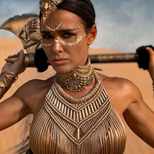 May include: A woman wearing a gold-colored halter top and a gold-colored headpiece with a large, ornate design. She is holding a large, gold-colored axe in her hand.