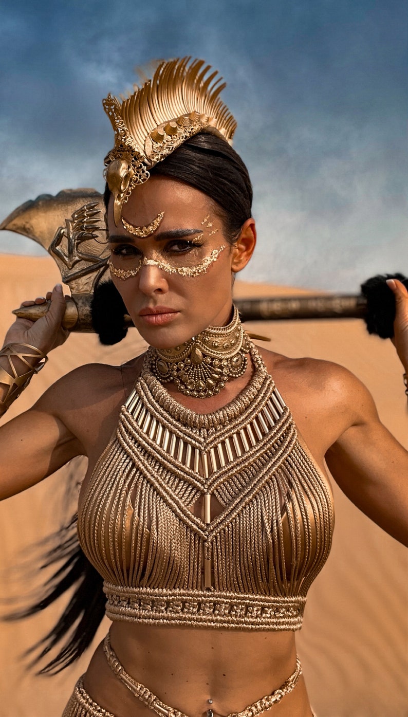 May include: A woman wearing a gold-colored halter top and a gold-colored headpiece with a large, ornate design. She is holding a large, gold-colored axe in her hand.