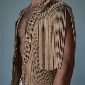 May include: A sleeveless garment made of woven gold-colored cords with fringe detailing. The garment is open in the front and has a V-neckline.
