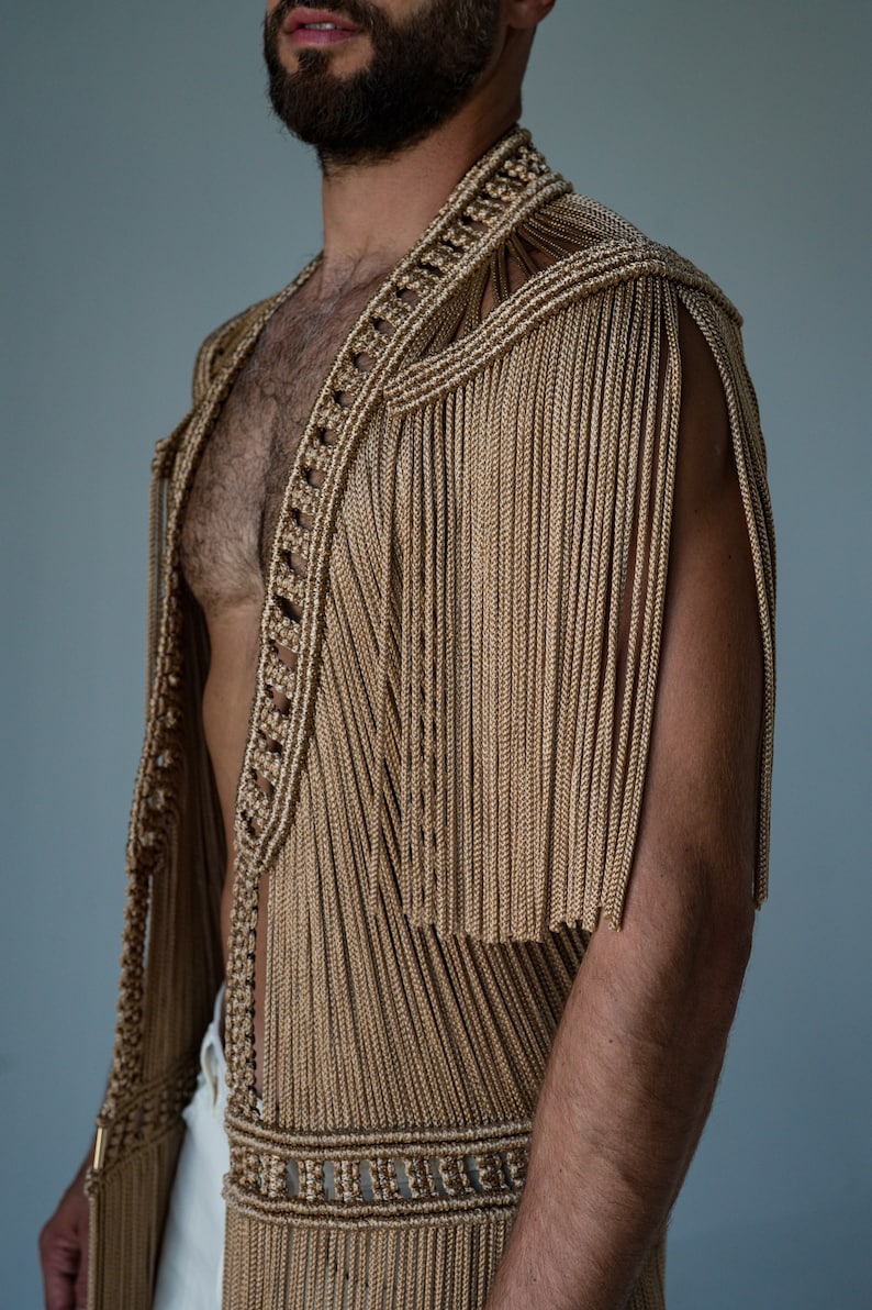 May include: A sleeveless garment made of woven gold-colored cords with fringe detailing. The garment is open in the front and has a V-neckline.