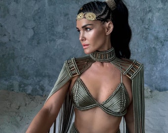 Puede incluir: Una mujer con un atuendo de crochet verde con detalles dorados. El atuendo incluye un top halter, una falda y una capa. La mujer lleva una diadema dorada con un diseño circular.