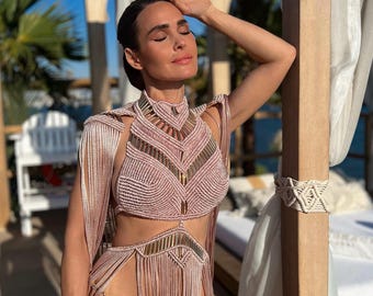 Puede incluir: Un conjunto de dos piezas de macramé rosa claro con detalles dorados. La parte superior tiene un cuello alto y flecos, y la parte inferior es una falda de cintura alta con flecos. El atuendo se usa al aire libre a la luz del sol.