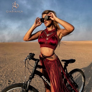 May include: A woman wearing a red crocheted top and skirt with fringe, standing next to a black bicycle with the word "Rockrail" on the frame. She is wearing goggles and has her hair pulled back.