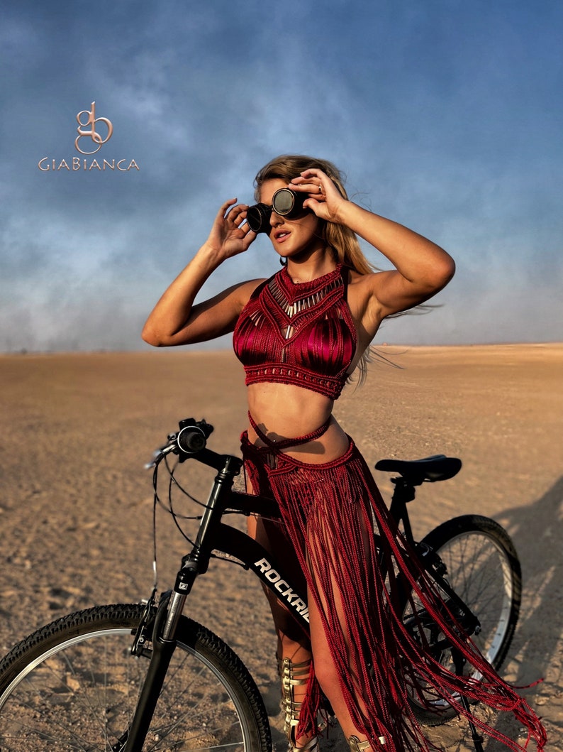 May include: A woman wearing a red crocheted top and skirt with fringe, standing next to a black bicycle with the word "Rockrail" on the frame. She is wearing goggles and has her hair pulled back.